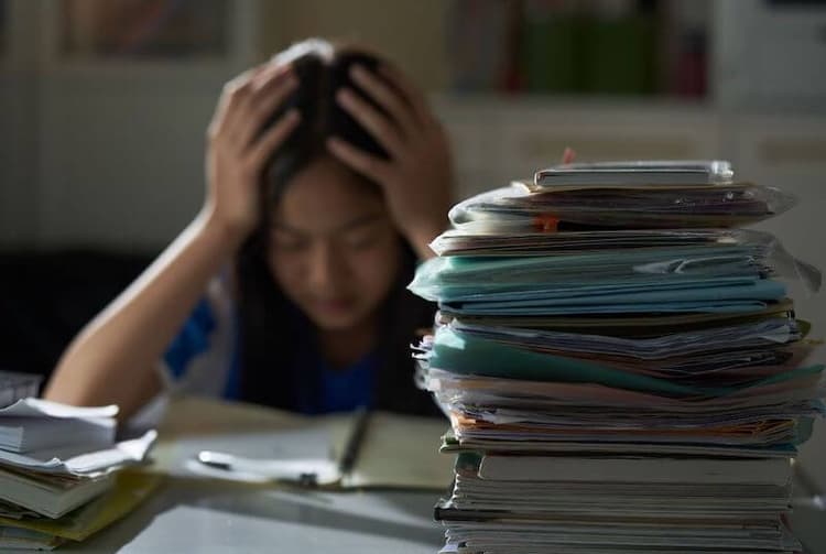 一名女子雙手抱頭伏在書桌前，桌上堆滿高疊的文件與檔案夾
