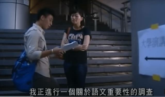 影片截圖:一名男生在校園樓梯前向一名女生遞上問卷,字幕為「我正進行一個關於語文重要性的調查」