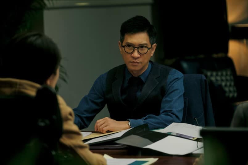 電影劇照:一名戴眼鏡的男子在會議桌前與對面的人對話,桌上放滿文件