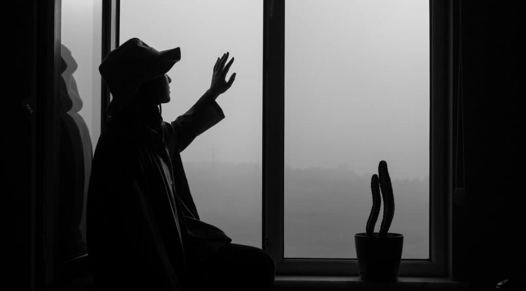 黑白剪影:一人戴帽坐在窗邊,抬手貼向玻璃望向窗外,窗台放著一盆仙人掌