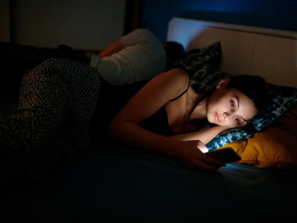 一名女子夜間側躺在床上看著發光的手機螢幕,身旁有人已入睡